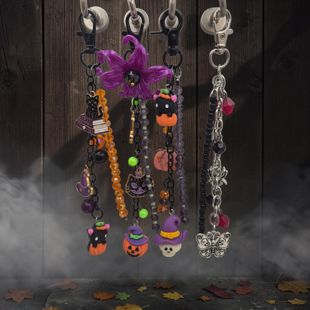 Four Halloween-themed beaded keychains hanging from silver lobster clasps against a dark wood plank background with mist and scattered autumn leaves. The keychains feature purple, orange, black, and silver beads and charms including a purple flower, witch hats, jack-o'-lanterns, a cat, a broom, a book, and a butterfly.