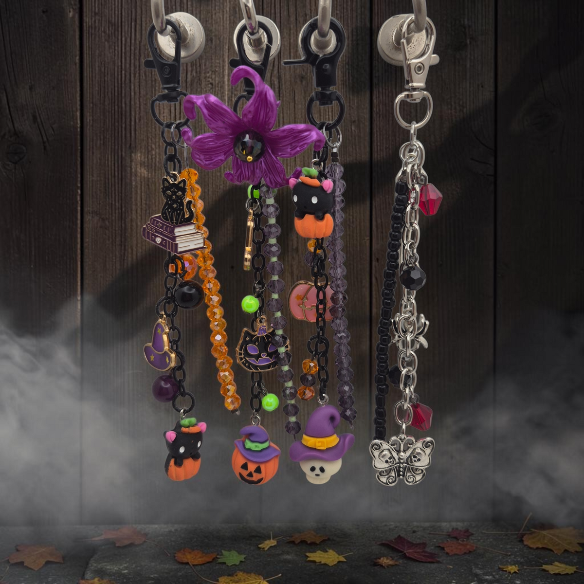 Four Halloween-themed beaded keychains hanging from silver lobster clasps against a dark wood plank background with mist and scattered autumn leaves. The keychains feature purple, orange, black, and silver beads and charms including a purple flower, witch hats, jack-o'-lanterns, a cat, a broom, a book, and a butterfly.